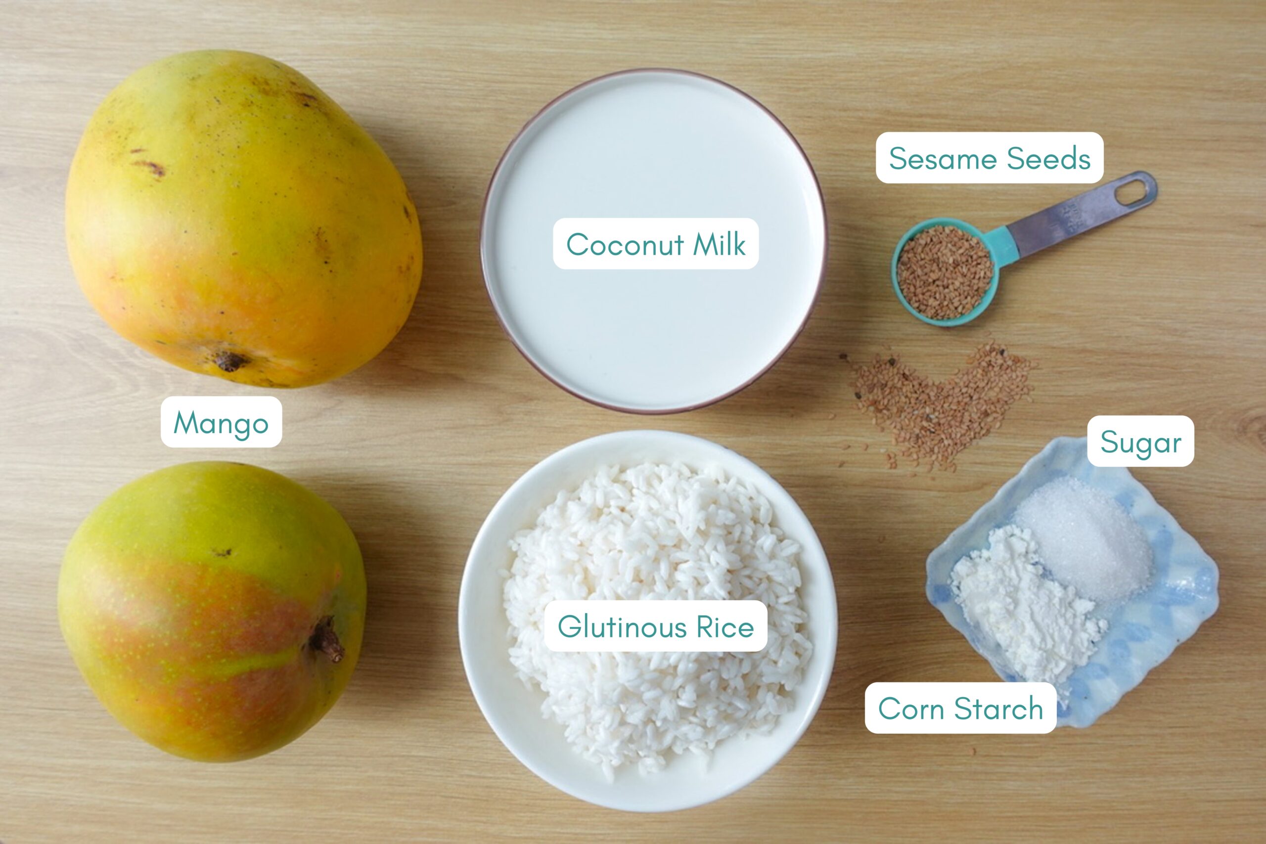 Ingredients laying out on a wooden table, including mango, coconut milk, glutinous rice, sesame seeds, sugar and corn starch.