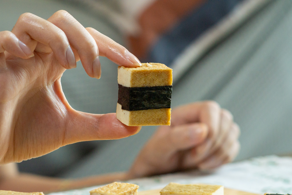 holding a piece of tofu wrapped by a strip of nori